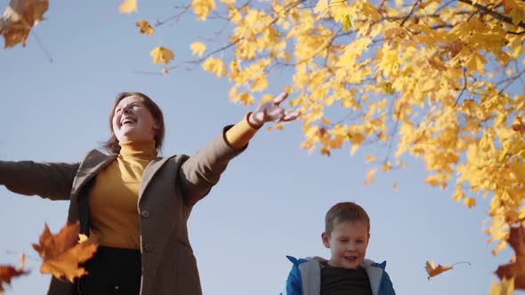 Happy Mom and Son Toss Up Yellow Leaves Against Blue Sky Autumn Yellowed Tree