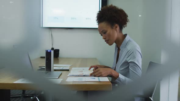 Mixed race businesswoman sitting at desk, checking documents and using laptop in office