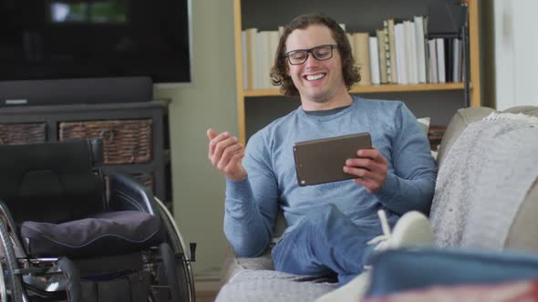 Happy caucasian disabled man sitting on sofa in living room using tablet