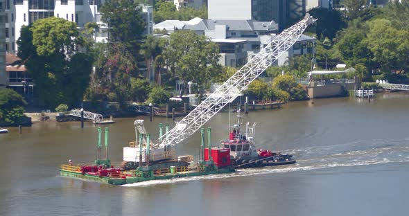 A small crane is transported up a river on a flat deck barge that is being manoeuvred by a small tug