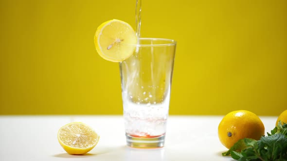 Glass with Lemon Slice at Yellow Background and Water Flowing Inside Spilling
