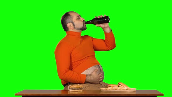 Overweight Caucasian Man Sits at a Table with Junk Food and Drinks Beer Stroking His Stomach, Green