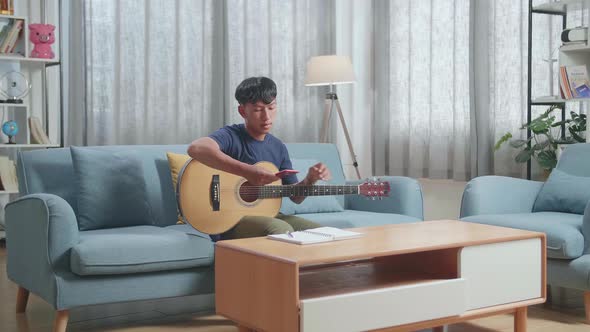 Asian Boy Composer With Note Book Looking At Smart Phone And Playing Guitar At Home