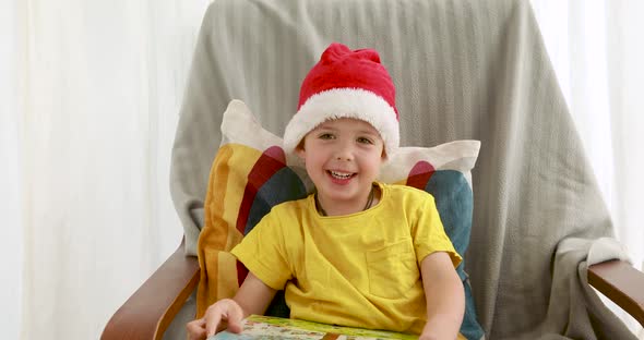 Funny Boy in Red White Christmas Hat Sits on Chair Smiling