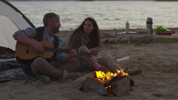 Man Playing Guitar and Woman Eating Sausage by Campfire on Beach