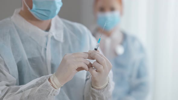 Closeup Hands of Unrecognizable Doctor Preparing Vaccine in Syringe with Blurred Nurse in Covid Face