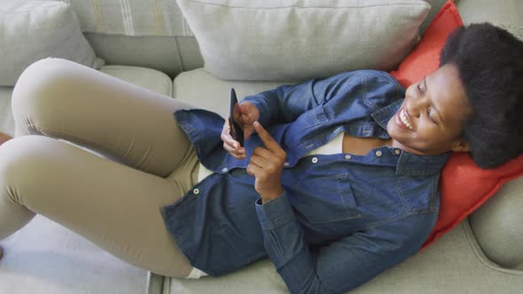 Happy african american woman laying on sofa, using smartphone