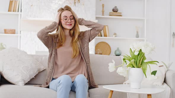 Young Adult Mother Blonde Woman in Glasses Sits on Sofa at Home Holding Head with Hands From Noisy