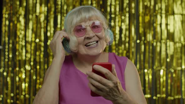 Stylish Happy Senior Old Woman Holding Cell Phone, Listening To Music in Headphones and Dancing