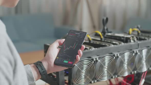 Man's Hand Holding Smartphone Showing Bitcoin Chart In Front Of Mining Rig For Mining Cryptocurrency