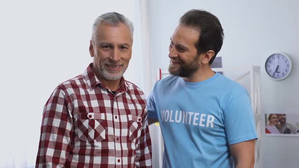 Young Male Volunteer Hugging Senior Male and Both Smiling to Camera, Charity