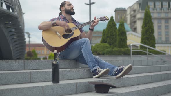 Wide Shot Portrait of Enthusiastic Musician Playing Guitar on City Stairs with Wine Bottle and Hat