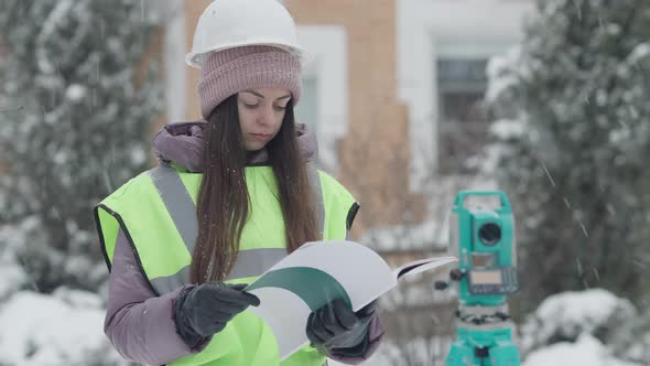 Concentrated Female Architect Examining Project Documentation on Snowy Winter Day Outdoors with