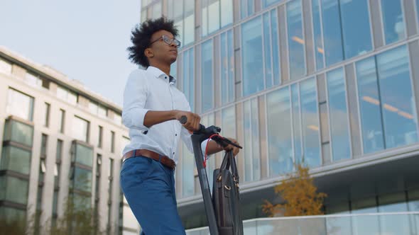 African Young Businessman in Glasses Riding Electro Scooter Near Office Building.