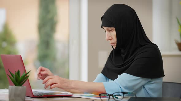 Side View Portrait of Intelligent Middle Eastern Mature Woman in Hijab Messaging Online on Laptop