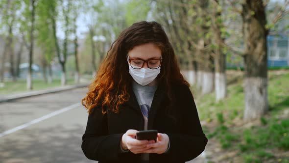 Young Girl in a Medical Mask Uses a Smartphone in the Park