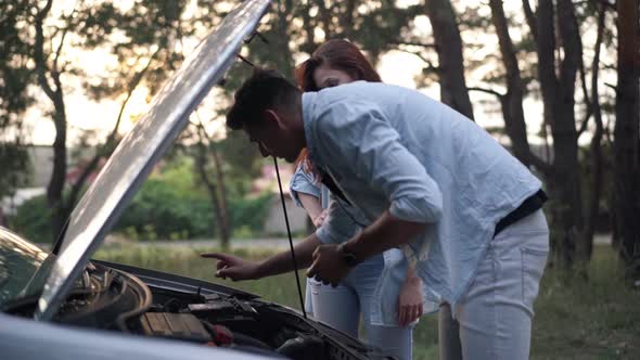 Side View Portrait of Focused Man Leaning at Open Car Hood Talking with Woman Standing at Background