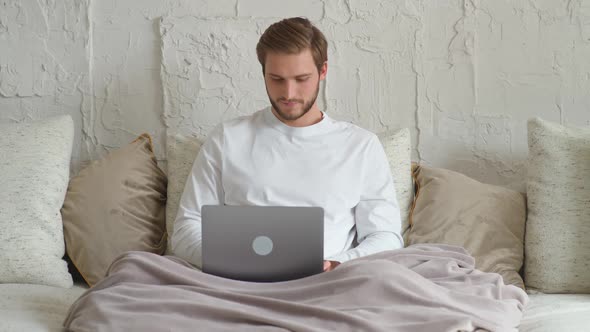 Cheerful Caucasian Man Sits at Home on the Couch and Works Using a Laptop Financial Manager on a