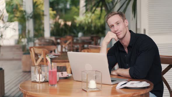 Man Freelancer Sitting at Table with Laptop in Modern Cafe, Looks Away, Laughing