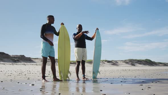 African american father and teenage son standing on a beach holding surfboards and talking