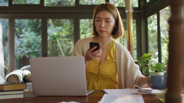 Asian woman sitting at table working from home and using smartphone