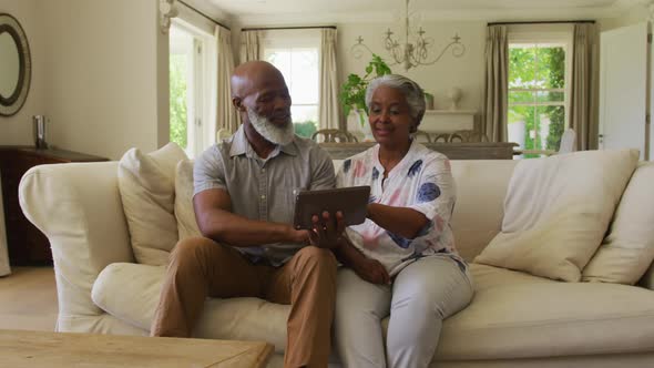 African american senior couple looking at each other and smiling while using digital tablet at home