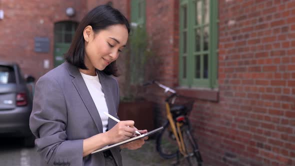 Happy Young Asian Woman Taking Notes Using on Digital Tablet Standing Outside