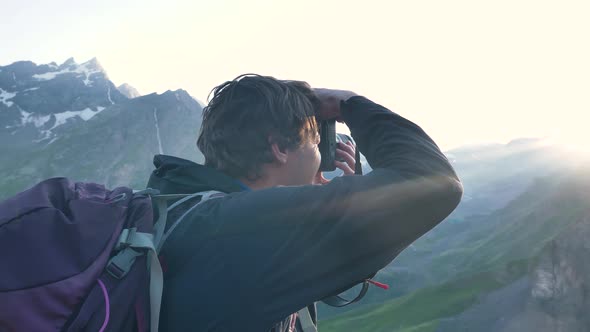 A Male Traveler with a Backpack Takes a Photo in the Mountains in the Rays of the Setting Sun While