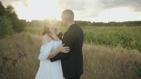 The Bride and Groom Stand Hugging on the Edge of a Corn Field in the Rays of the Sun Breaking
