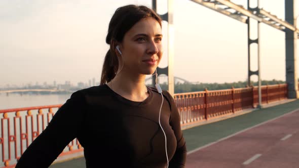 Portrait of a Pretty Confident Woman with Black Sportswear and White Earphones on the Bridge