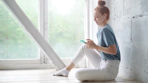 Happy Redhead Young Woman Wearing Wireless Earphones Is Using Cell Phone Sitting By the Big Window