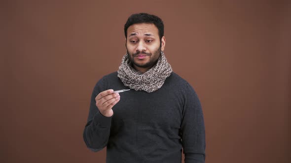 Annoyed Indian Man in Scarf Looking at Thermometer Feeling Unwell and Sick Brown Studio Background