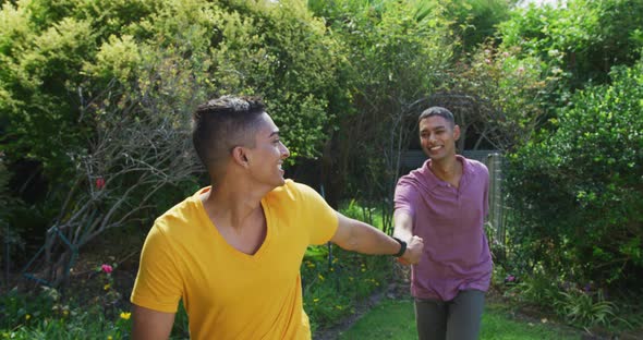 Smiling mixed race gay male couple holding hands walking in garden