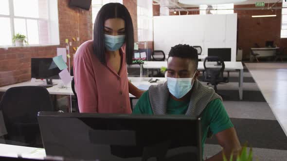 Diverse male and female business colleagues wearing face masks in discussion at work using computer