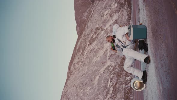 Astronaut Without a Helmet in a Chair By the Hills Drinking From Bottle