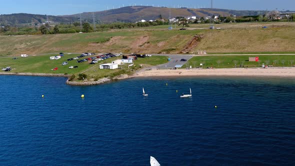 people practicing in the sailing school of the lake with beach, car parking and wind turbines in the