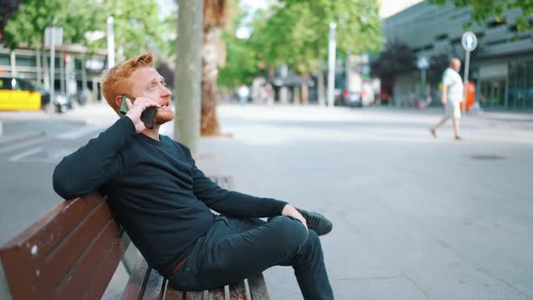 Serious ginger man talking by smartphone while sitting on the bench