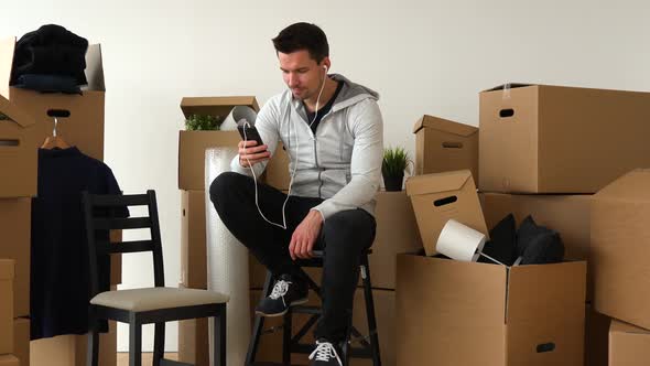 A Moving Man Sits on a Chair in an Empty Apartment and Listens To Music on a Smartphone