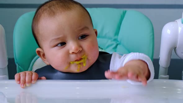 A dirty, joyful baby at the table on a feeding chair got his mouth dirty in vegetable puree and knoc