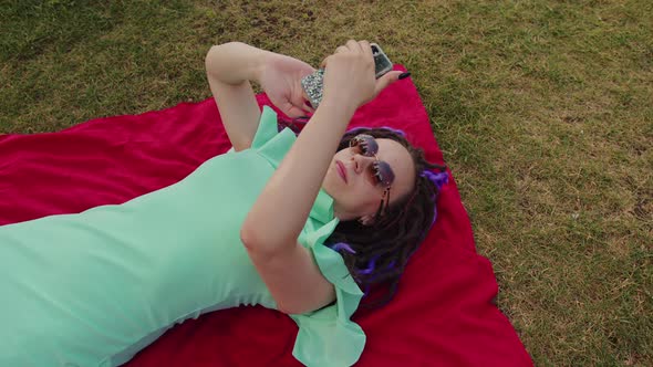 Young woman lying on a blanket blowing uses a smartphone in the park.