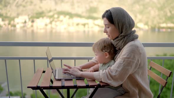 Mother Multitasking Holding Baby Infant and Using Computer Laptop at Home