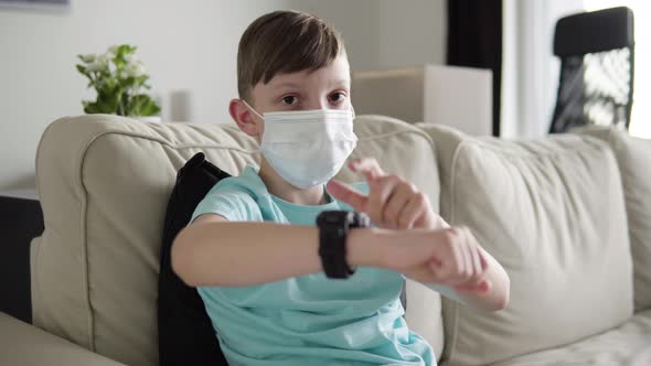 A Young Boy in a Face Mask Taps His Watch at the Camera and Nods As He Sits on a Couch at Home