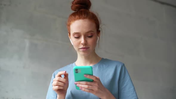 Cheerful Redhead Young Woman Wearing Wireless Earphones Is Listening Pleasant Music on Cellphone