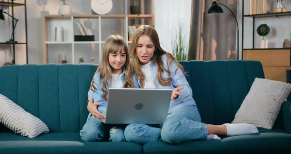 Friendly Two Blond Sisters Sitting on Soft Couch at Home During Positive Video Call on Laptop