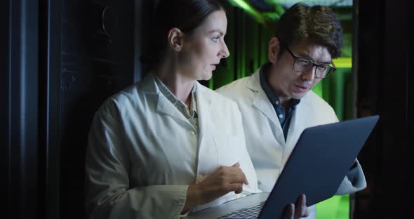 Diverse female and male it technicians in lab coats using laptop checking computer server