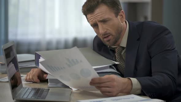 Man Surrounded by Paperwork at Office Looking at Graphs, Head Falling on Folders