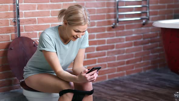 Domestic Young Woman Smiling Using Smartphone Sitting on Toilet at Home Bathroom