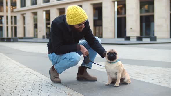 Young Hipster Man Squatting and Stroking Cute Pug Dog Outdoors