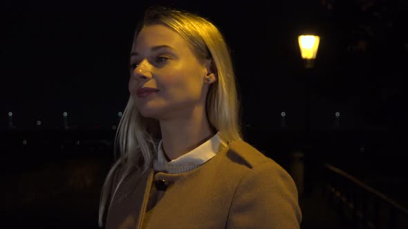 A Young Beautiful Woman Looks Around with a Smile in an Urban Area at Night