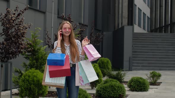 Positive Young Girl with Bags Talking on Mobile Phone About Sales in Shopping Mall in Black Friday